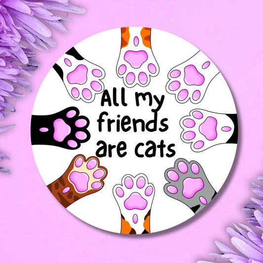 A round pin badge featuring the text 'All my friends are cats' surrounded by illustrations of various cat paws in different colours on a white background, styled on a purple background with purple flowers in the corners.