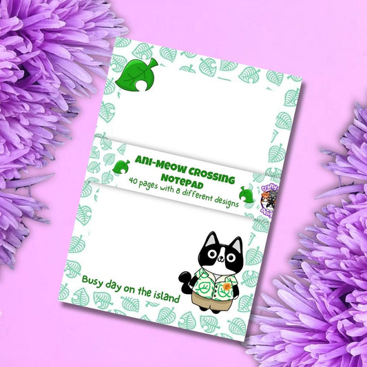 A notepad featuring the 'Ani-Mew Crossing' theme, styled flat-lay on a pink background with purple flowers. The notepad has a white base with a green leaf pattern border, a central white band with green text, and cartoon illustrations of a black cat in a green shirt and a green dog.