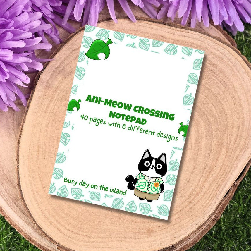 Notepad with cat design on a wooden surface with purple flowers. "8 unique page designs" and "handmade in small batches