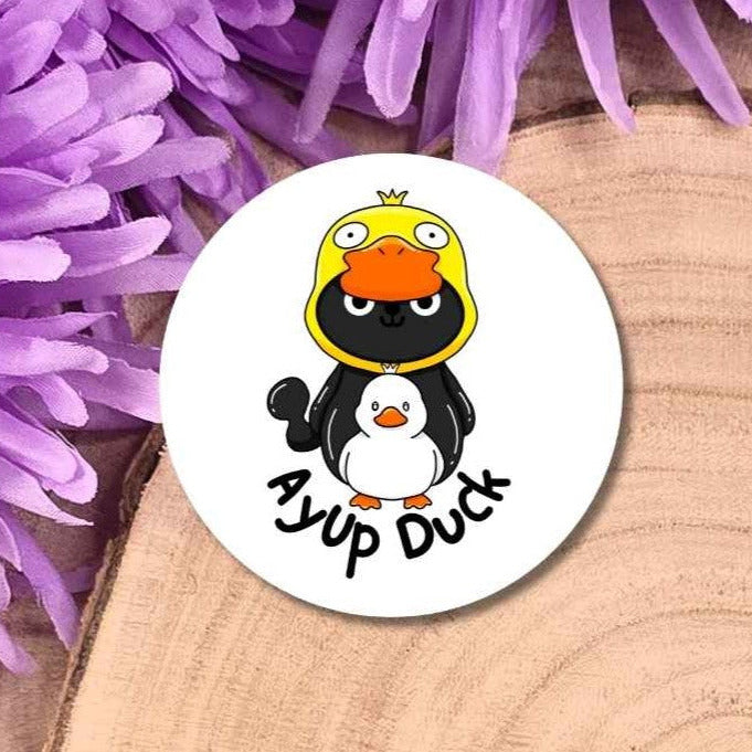A round white badge featuring a cartoon cat wearing a yellow duck hood with an orange beak, placed on a wooden surface with purple flowers around it. The sticker reads 'Aup Duck'.