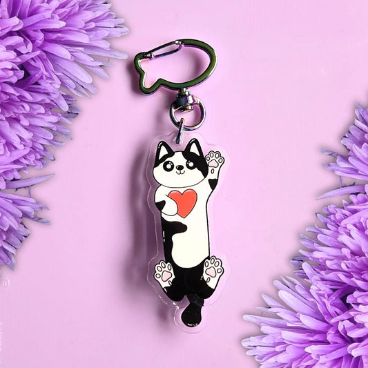 front view of a black and white cow print cat keyring with moveable tail and fish shaped keyring attachment. laid on a purple background with purple flowers