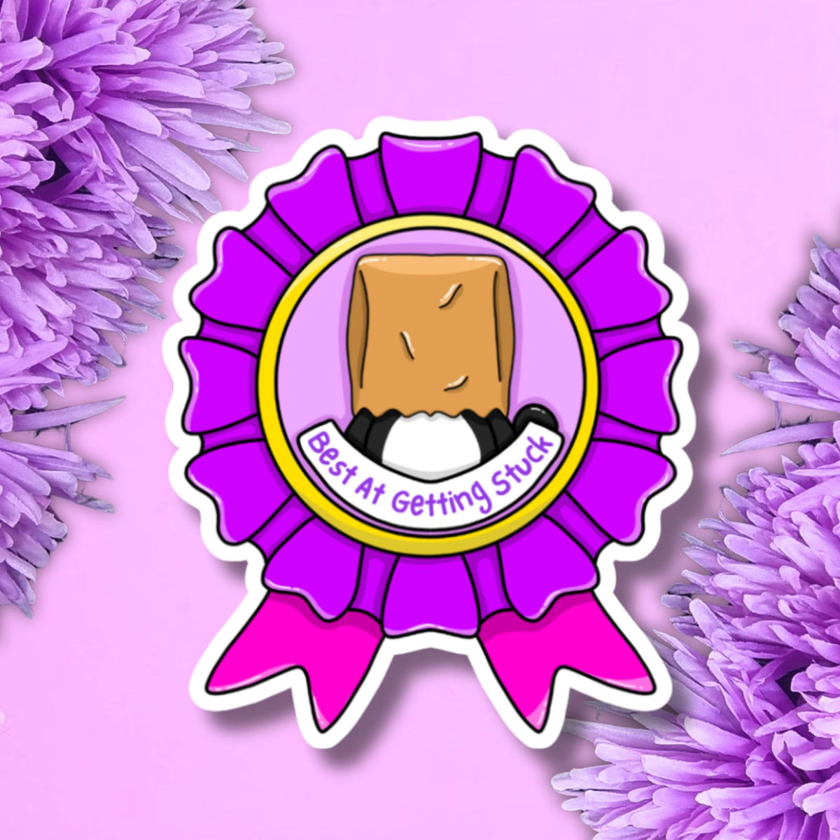 A die-cut sticker featuring a purple ribbon badge with a cartoon brown paper bag character and the text 'East At Getting Stuck', shown on a pink background with purple flowers.