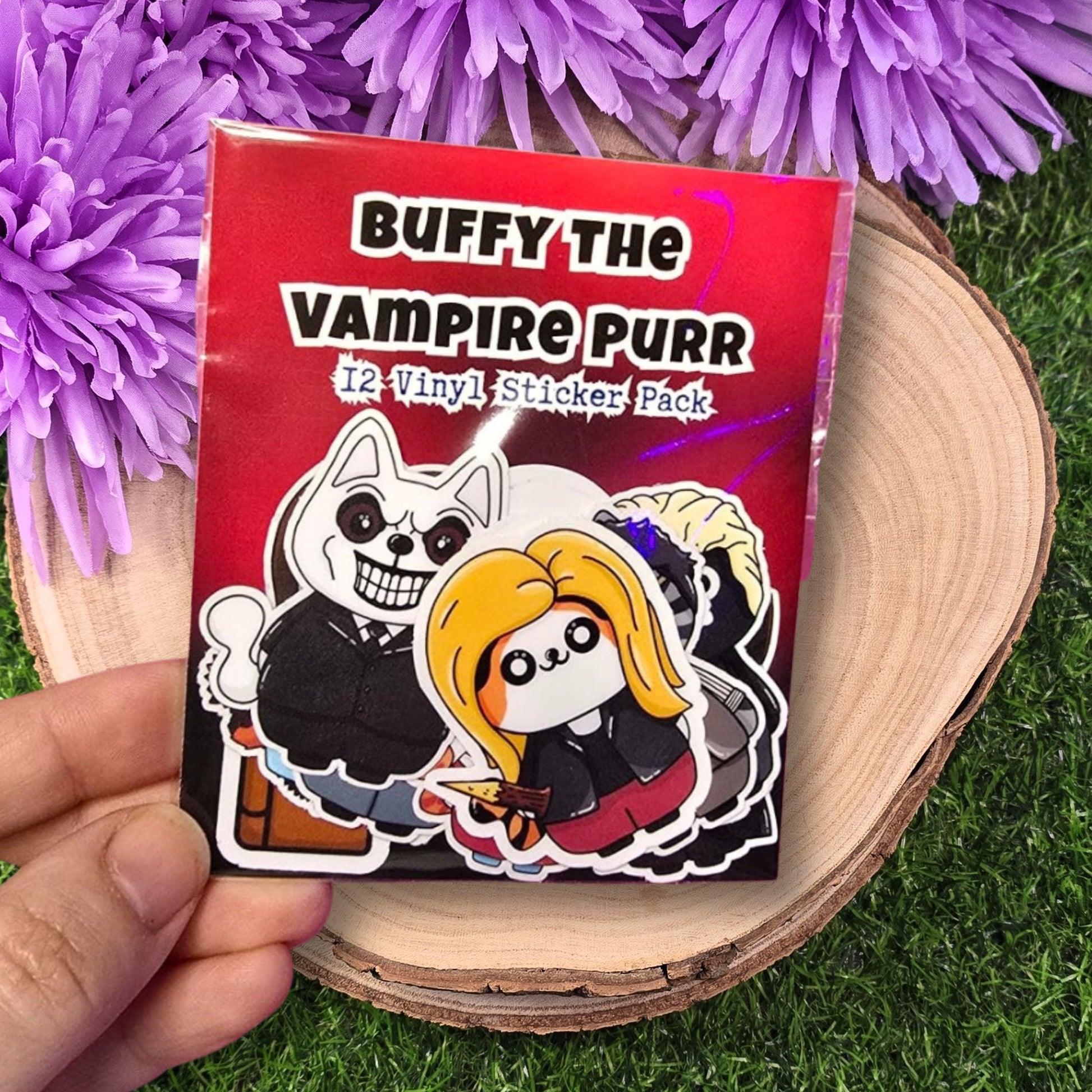 Pack of 'Buffy the Vampire Purr' vinyl stickers held over a wooden surface with purple flowers in the background.