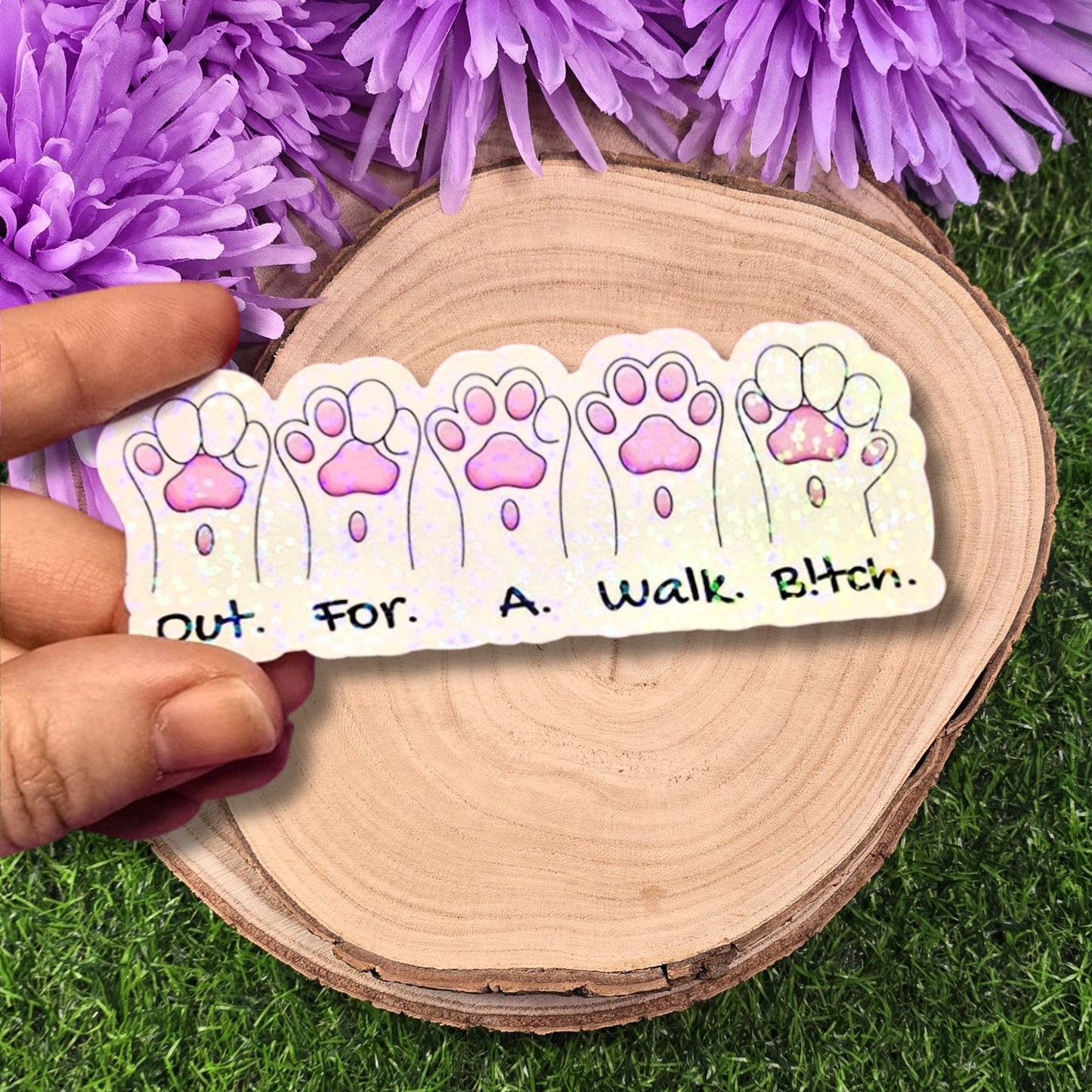 Sticker with paw prints and text held over a wooden surface with purple flowers in the background