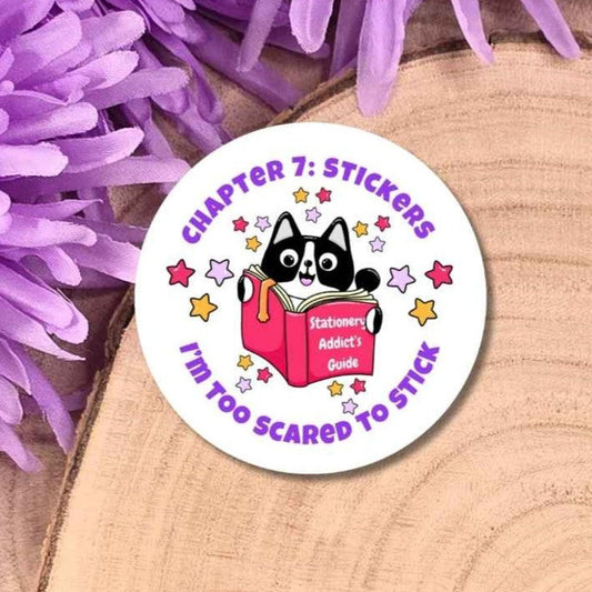 A round sticker featuring a black and white cat reading a pink book with purple text, displayed on a wooden slice with purple flowers.