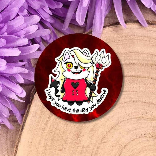 A round pin badge featuring a cartoon devil cat character in a red dress with the text 'I hope you have the day you believe,' displayed on a wooden slice with purple flowers in the background.