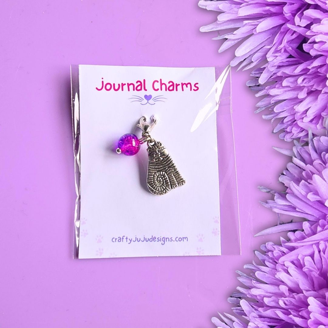 A silver-toned journal charm featuring a dangling cat pendant and a small bright pink purple bead, displayed on a white card labeled 'Journal Charms' against a purple background with purple flowers at the side.