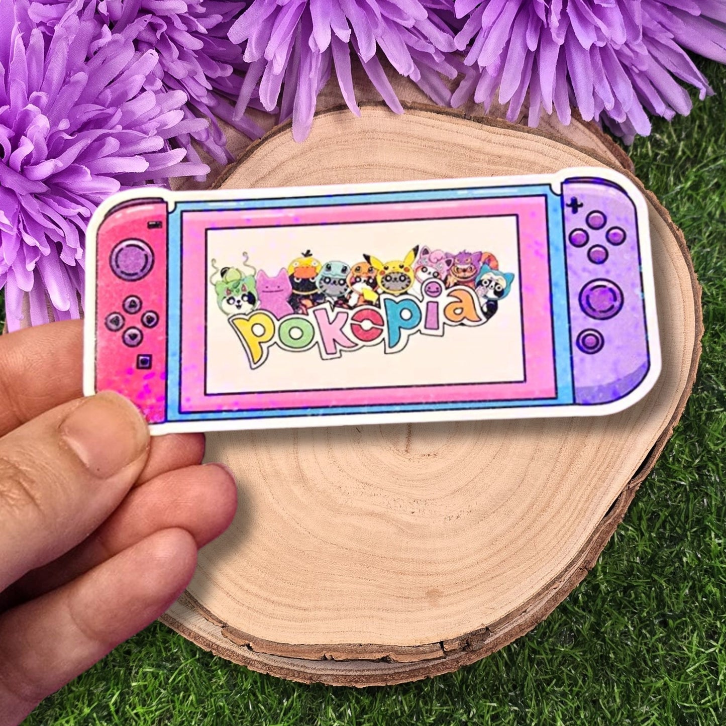 Sticker of a colorful gaming console with 'Pokopia' branding held over purple flowers on grass.