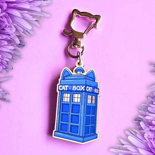 A blue TARDIS-shaped keychain charm with a gold-tone clasp, photographed on a pink background with purple flowers framing the sides.