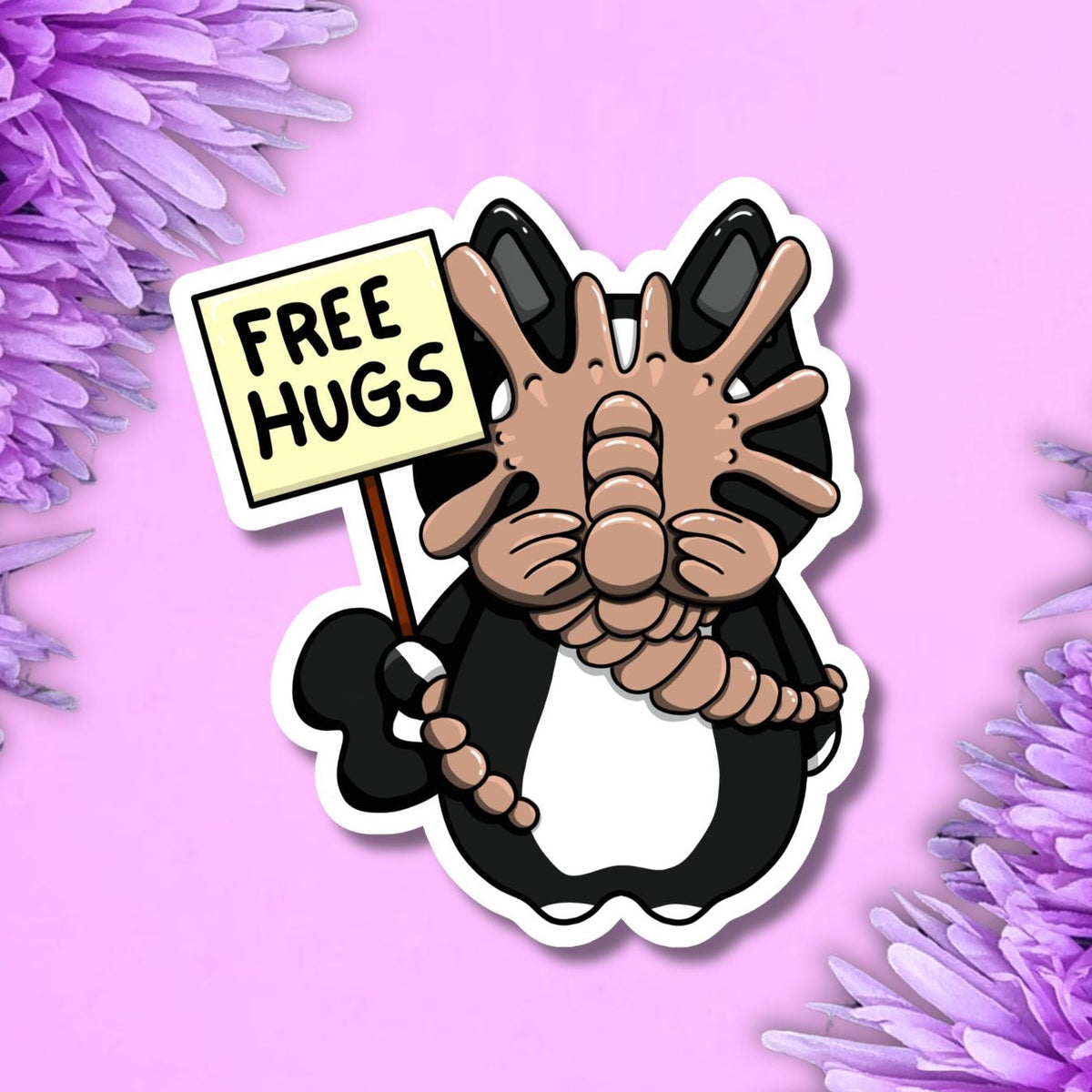 A die-cut sticker featuring a cartoon penguin holding a sign that reads 'FREE HUGS' in black text on a cream background. The sticker is shown on a light pink background with purple flowers framing the corners.