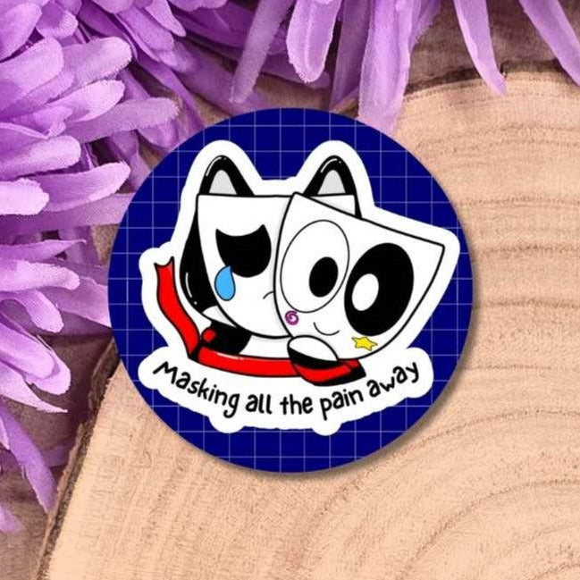 A round sticker featuring a cartoon cat character with a teardrop and the text 'Masking all the pain away', placed on a wooden surface with purple flowers in the background.