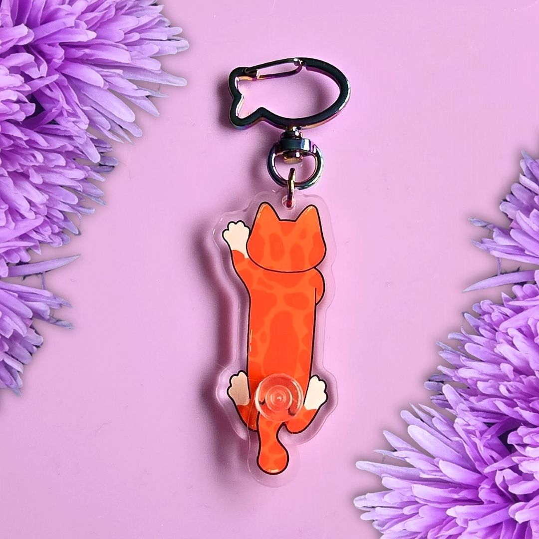 An ginger cat keychain charm with a fish shaped metal clip, displayed on a purple background with purple flowers in the corners.