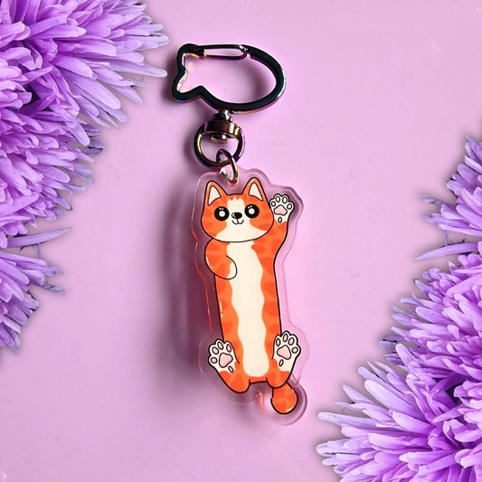 A cute ginger cat key chain charm with a fish-shaped clip, displayed on a purple background with purple flowers in the corners.