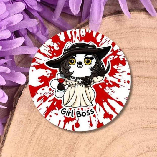 A round pin badge featuring a cartoon cat in a hat with the text 'Girl Boss' on a red and white splatter background, displayed on a wooden slice with purple flowers.