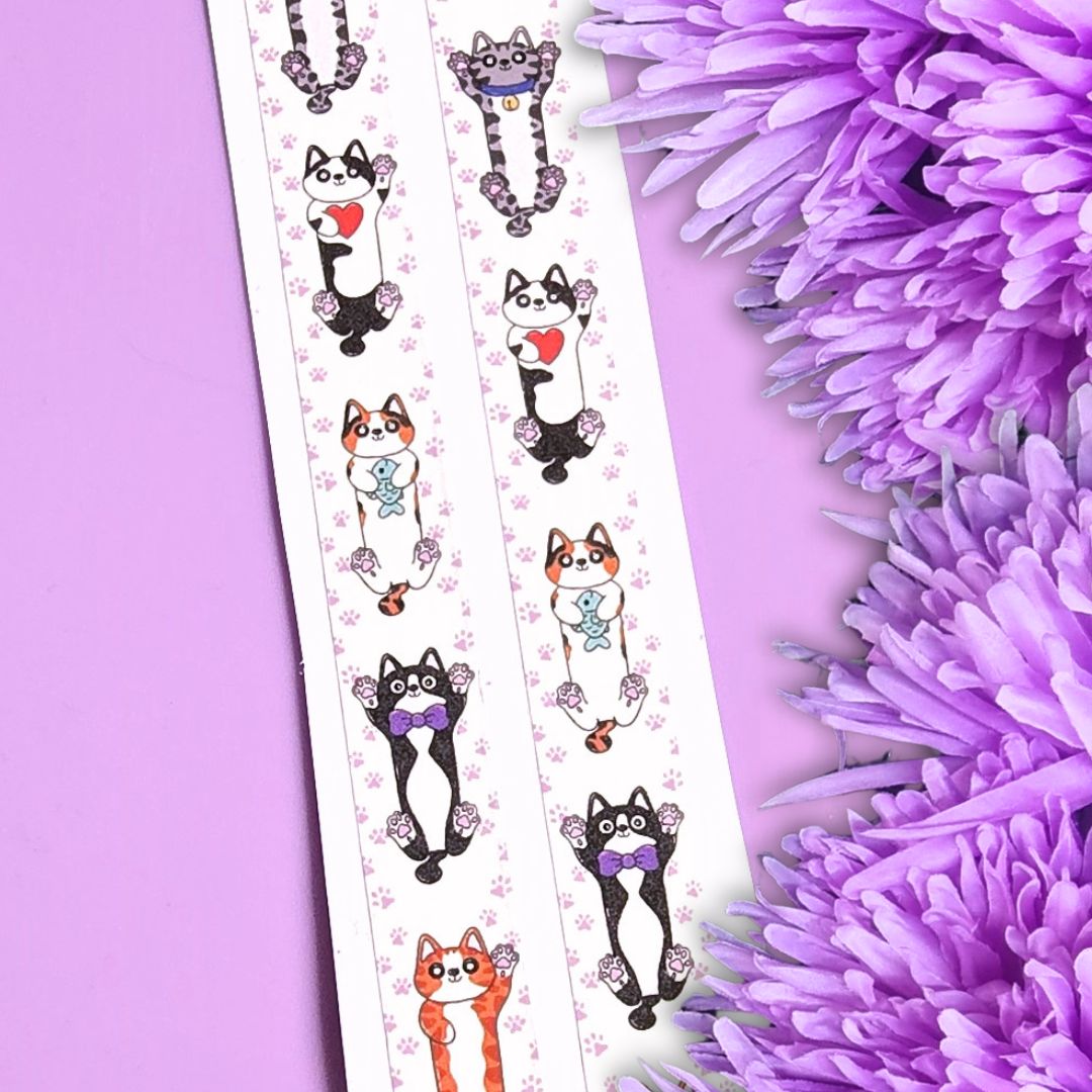 A decorative strip of washi tape featuring cute illustrated cats in black, white, and orange with purple bows and floral motifs, laid diagonally on a lavender background with purple flowers at the side.