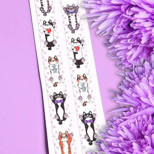 A decorative strip of washi tape featuring cute illustrated cats in black, white, and orange with purple bows and floral motifs, laid diagonally on a lavender background with purple flowers at the side.