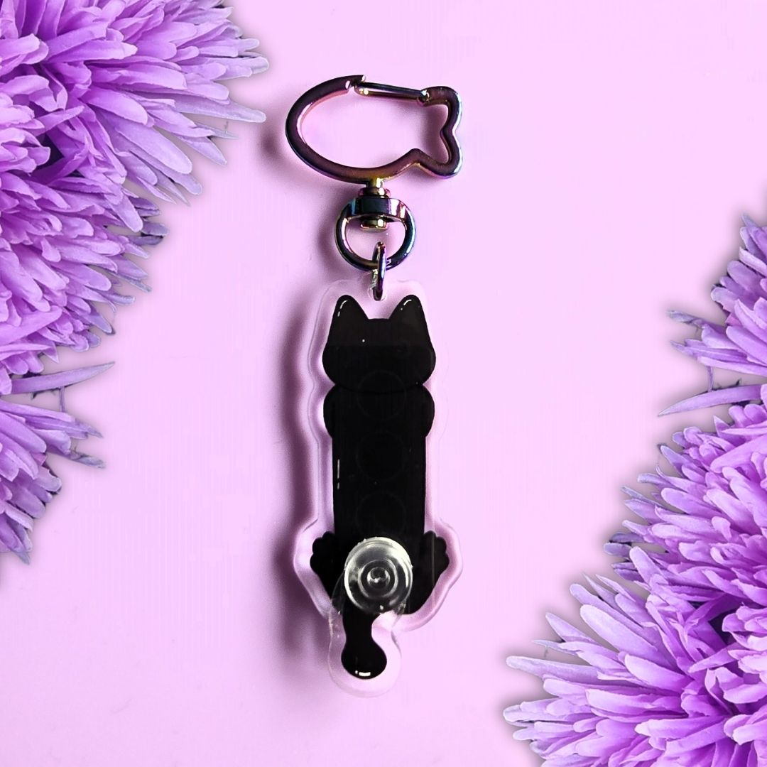 A black cat-shaped key chain with a metal clasp and key ring, displayed on a light pink background with purple flowers framing the sides.