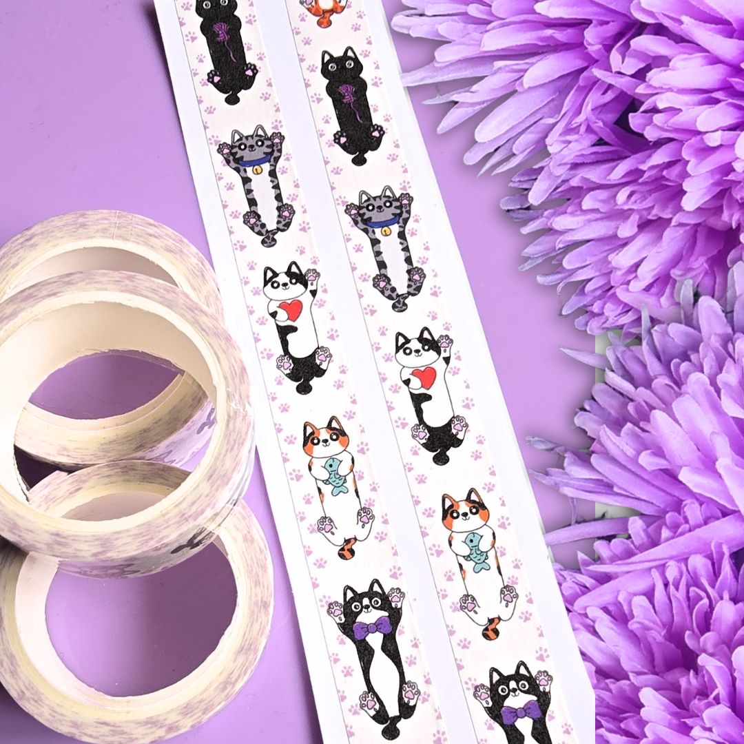 A roll of decorative washi tape featuring cute cartoon cats in black, white, purple, and orange, shown alongside two rolls of plain tape on a purple background with purple flowers.