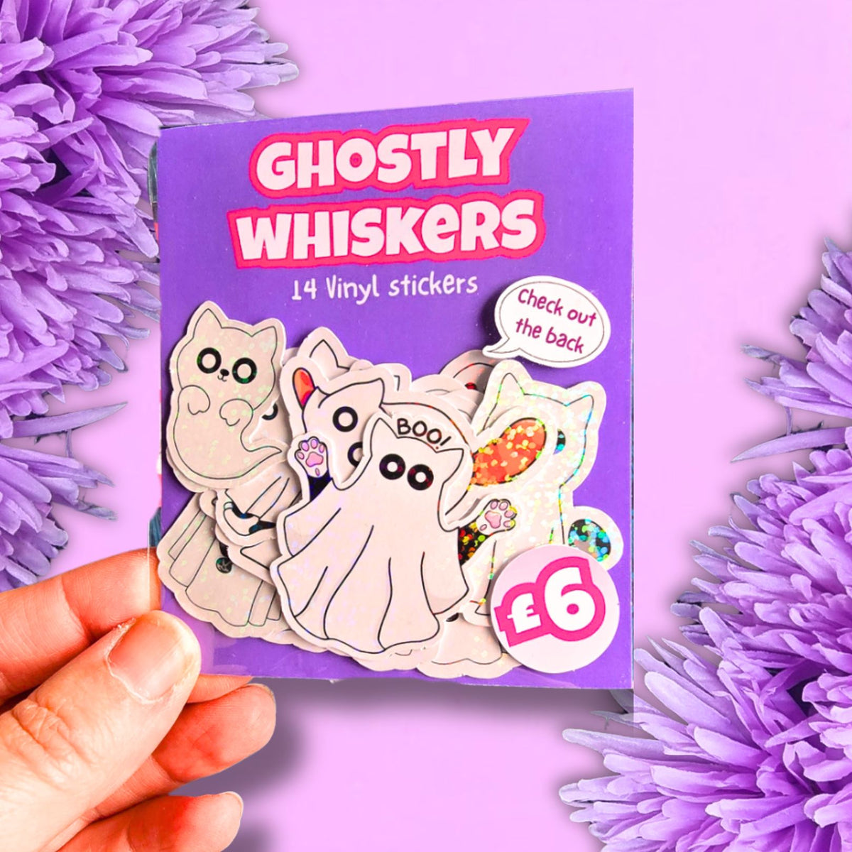 A hand holds a purple sticker pack displaying 'GHOSTLY WHISKERS' vinyl stickers, surrounded by purple flowers. The card shows several ghost-themed stickers, including one with whiskers and a ghost with a bow tie.