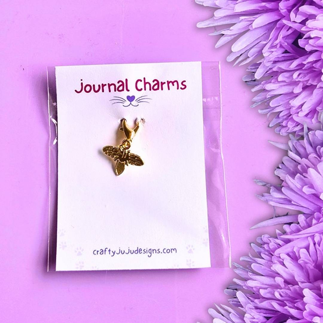 A small gold bee journal charm on a white display card labeled 'Journal Charms' by craftyJuJudesigns.com, photographed on a lavender background with purple flowers along the right edge.