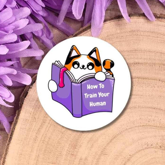 A round badge featuring a cartoon cat reading a purple book with the text 'How To Train Your Human', placed on a wood slice background with purple flowers.