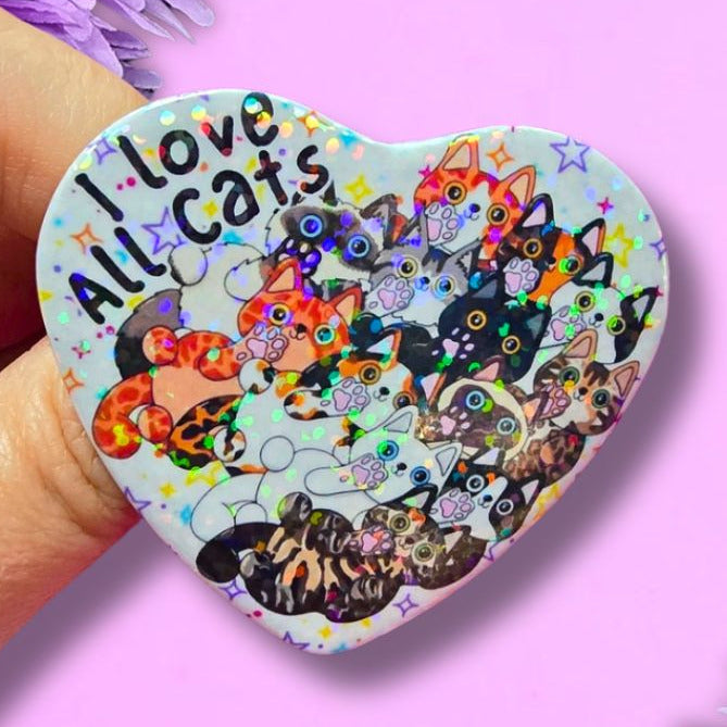 A hand holds a heart-shaped pin featuring a colorful illustration of various cats and the text 'I love All Cats' against a light background. The pin is set against a pink background decorated with purple flowers.