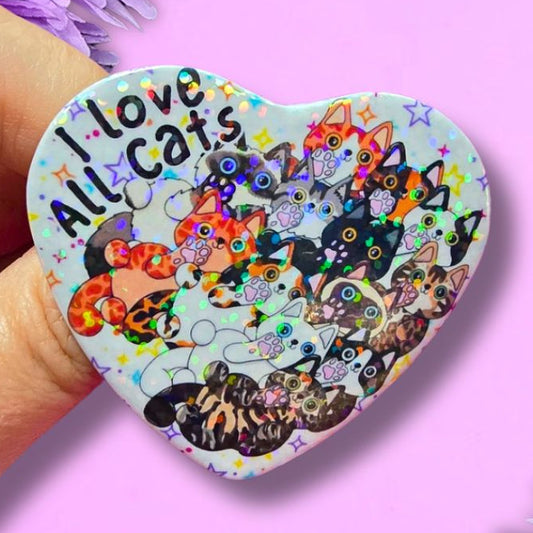 A hand holds a heart-shaped pin featuring a colorful illustration of various cats and the text 'I love All Cats' against a light background. The pin is set against a pink background decorated with purple flowers.