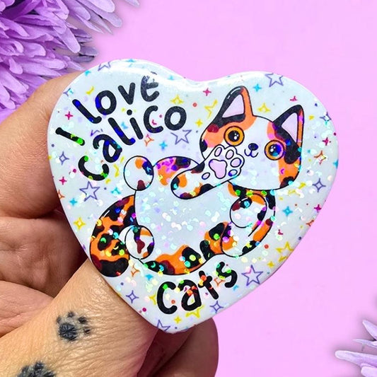 A hand holds a heart-shaped pinback button featuring a colourful cartoon cat and the text 'I love Calico cats' against a purple background with purple flowers.