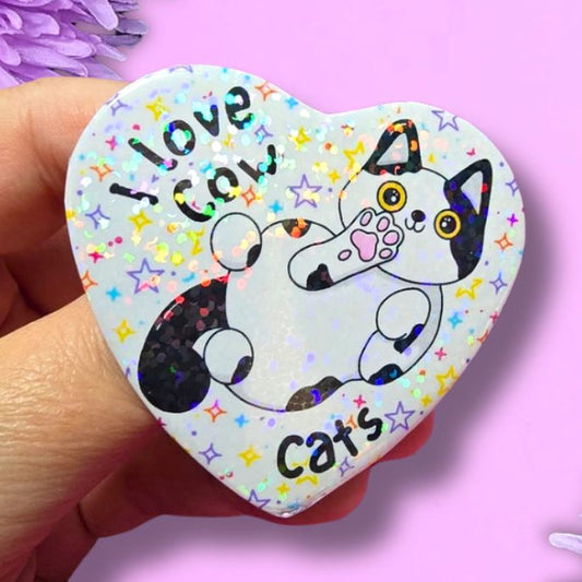 A hand holds a heart-shaped pin featuring a black and white cat illustration with the text 'I love cats' on a glittery white background. The pin is set against a pink background decorated with purple flowers.