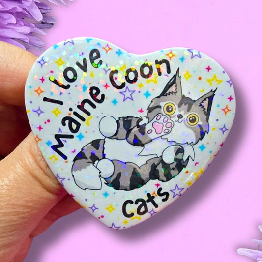 A hand holds a heart-shaped pin badge featuring a Maine Coon cat illustration and the text 'I Love Maine Coon Cats' against a pink background with purple flowers.