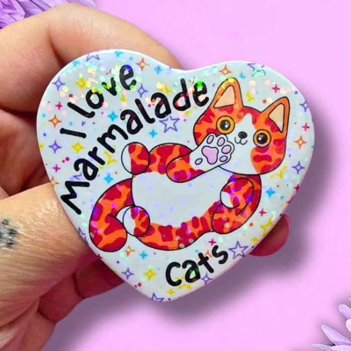 A hand holds a heart-shaped pin badge featuring a colorful cartoon cat and the text 'I love Marmalade Cats' against a pink background with purple flowers.
