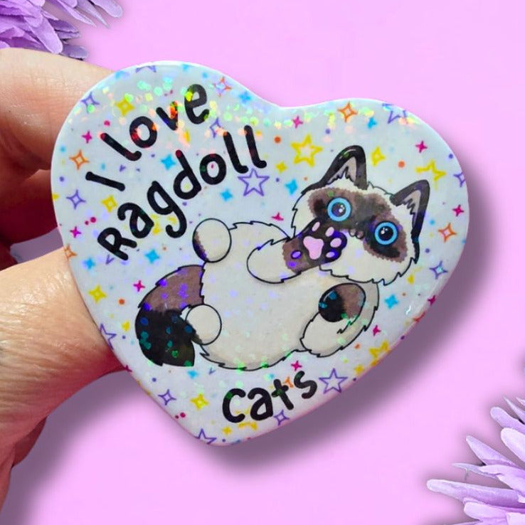 A hand holds a heart-shaped pin featuring a cartoon cat illustration and the text 'Love Ragdoll Cats' against a pink background with purple flowers.