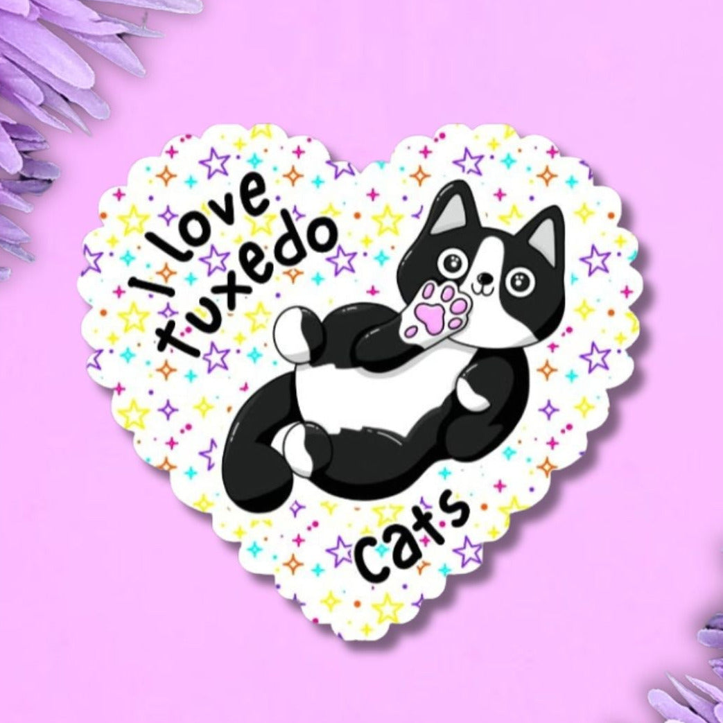 A heart-shaped sticker featuring a black and white tuxedo cat illustration with the text 'I love tuxedo cats' and small colourful stars, displayed on a purple background with purple flowers in the corners.