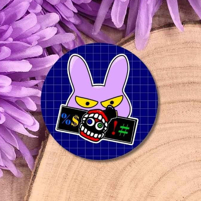 A round pin badge featuring a cartoon rabbit character with yellow eyes, set against a dark blue grid background. The badge is displayed on a light wood surface next to purple flowers.