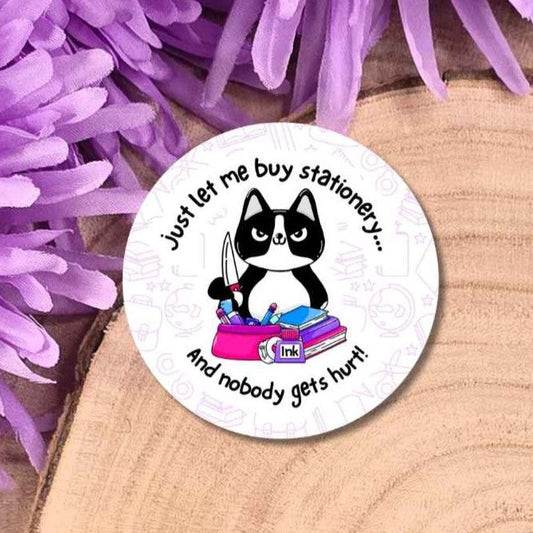 A round badge featuring a black-and-white cat sitting behind stationery holding a knife and the text 'just let me buy stationery.