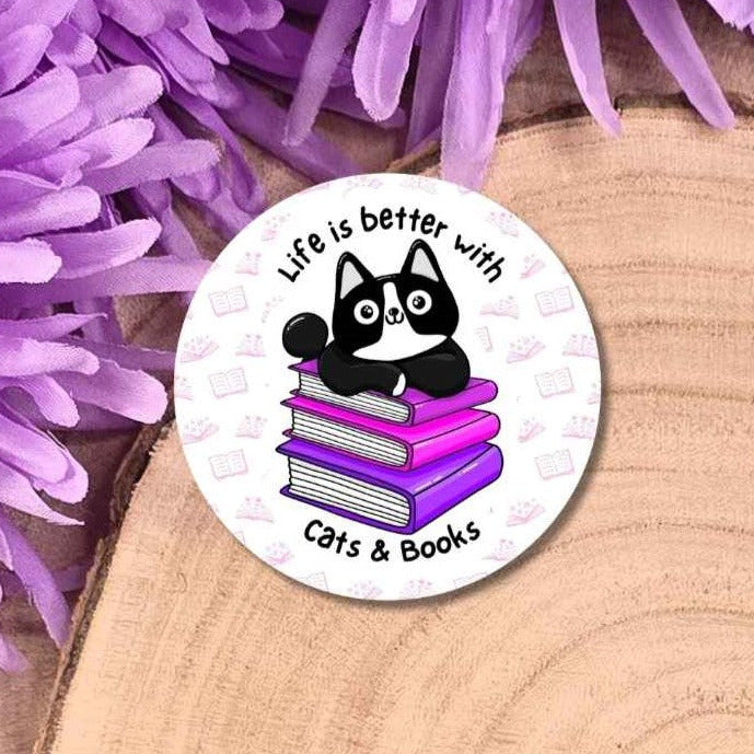 A round white pin badge featuring a black and white cat sitting on a stack of purple books with the text 'Life is better with Cats & Books', displayed on a wooden slice with purple flowers in the background.