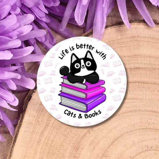 A round white pin badge featuring a black and white cat sitting on a stack of purple books with the text 'Life is better with Cats & Books', displayed on a wooden slice with purple flowers in the background.