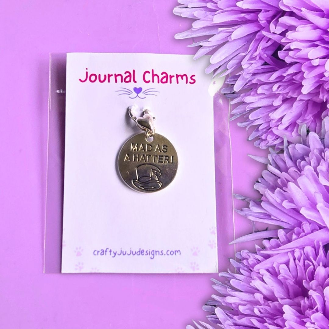 A round metallic charm card with the engraved text 'MAD AS A HATTER!' displayed on a white backing card labeled 'Journal Charms' and 'craftyJuJudeSigns.com', photographed on a purple background with purple flowers at the side.