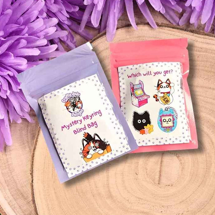 Two small mystery bags placed on a round wood slice with purple flowers around them. One bag is light purple with a white label reading 'Mystery keyring blind bag,' and the other is pink with a label reading 'which will you go?' and cartoon illustrations.