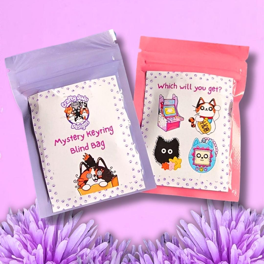 Two sealed mystery keyring blind bags displayed on a pink background with purple flowers at the bottom. One bag is lavender and the other is coral pink, both with white labels featuring cartoon cat illustrations and the text 'Mystery Keyring Blind Bag' and 'Which will you get?'.