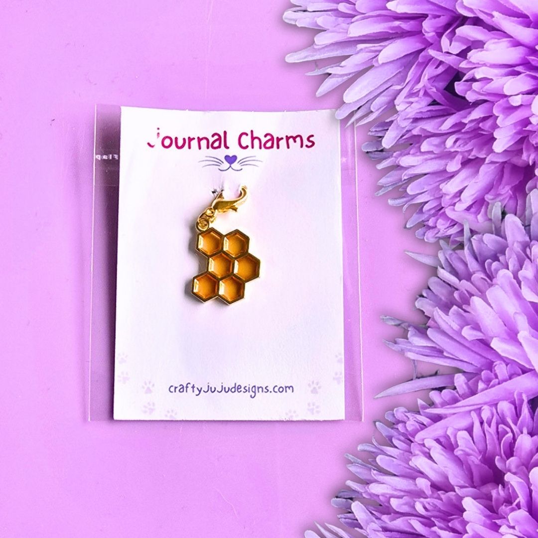 A honeycomb-shaped gold charm with a small butterfly detail, displayed on a white card labeled 'Journal Charms' and photographed on a pink background with purple flowers.