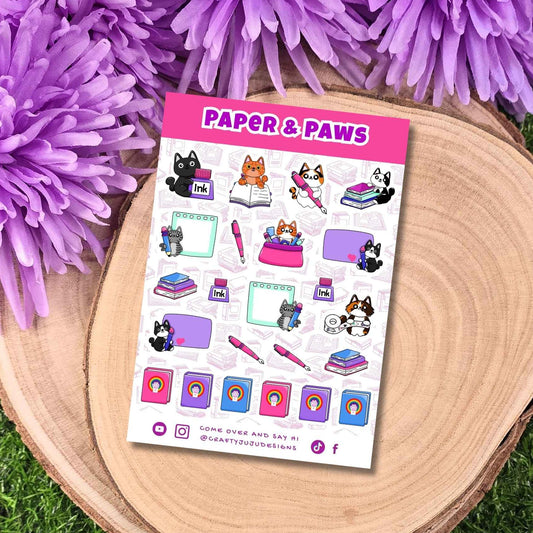 A sticker sheet titled 'PAPER & PAWS' featuring cute cat illustrations, placed on a round wooden slice with purple flowers around it.