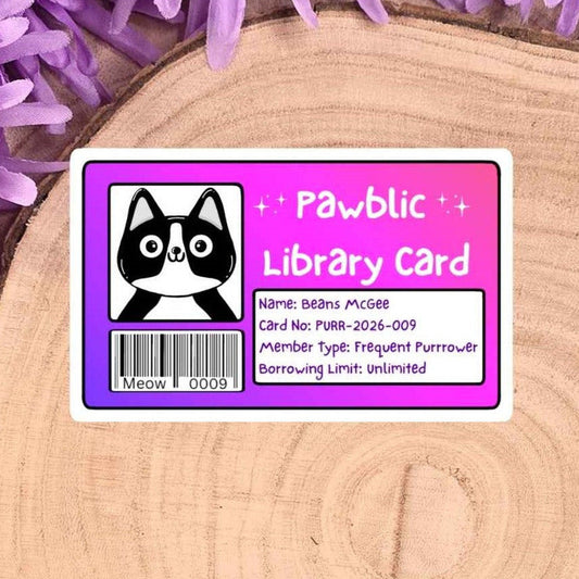 A rectangular library card sticker with a pink border and a black-and-white cat illustration, placed on a round wood slice with purple flowers around it.