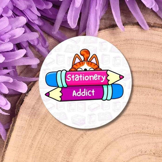 A round badge featuring a cartoon orange cat and two colourful pencils with the words 'Stationery Addict' on a white background, styled on a wood slice with purple flowers.