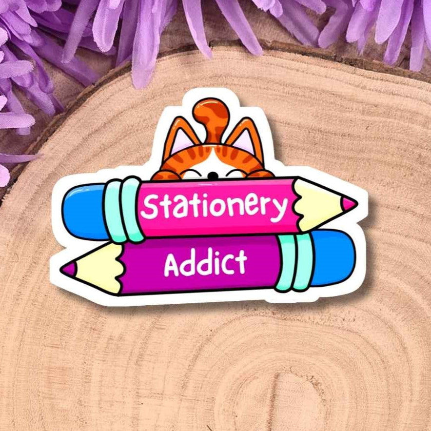 A die-cut sticker featuring a cartoon cat peeking over two colorful pencils with the words 'Stationery Addict' on them, placed on a round wood slice with purple flowers around it.