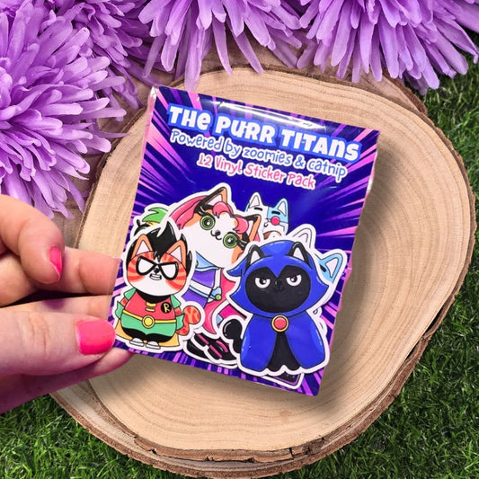 Pack of 'The Purr Titans' vinyl stickers held by a hand with pink nail polish, against a purple flower and wooden surface background.