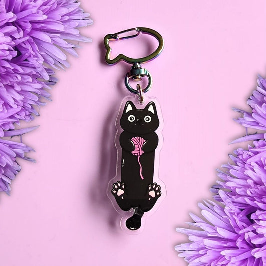 A black cat-shaped acrylic keychain featuring a pink yarn ball and white paw prints, attached to a silver metal keyring on a pink background with purple flowers.