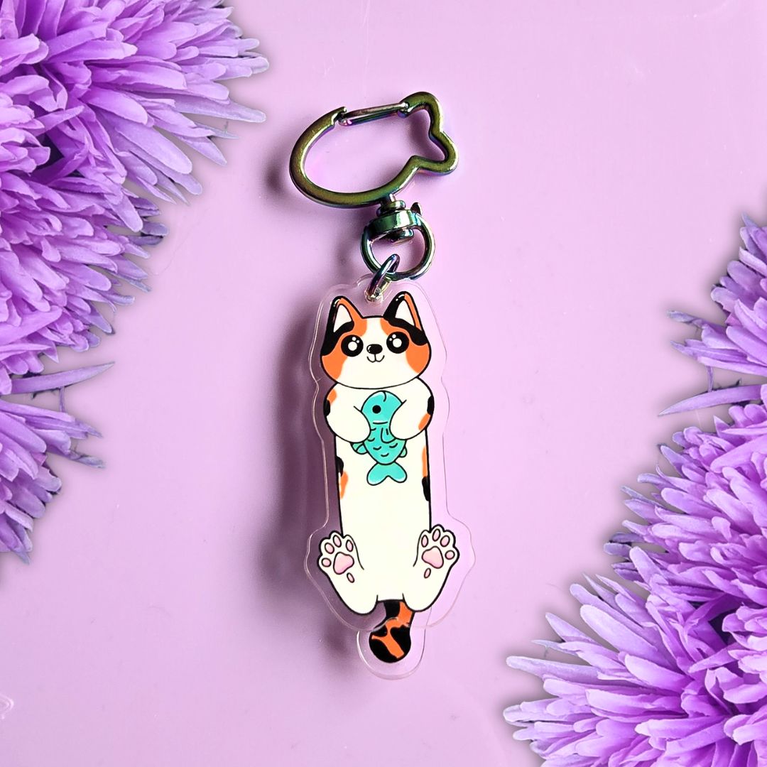 A cat keychain charm holding a blue fish, attached to a metal key ring, displayed on a light purple background with decorative purple flowers in the corners.