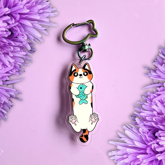 A cat keychain charm holding a blue fish, attached to a metal key ring, displayed on a light purple background with decorative purple flowers in the corners.
