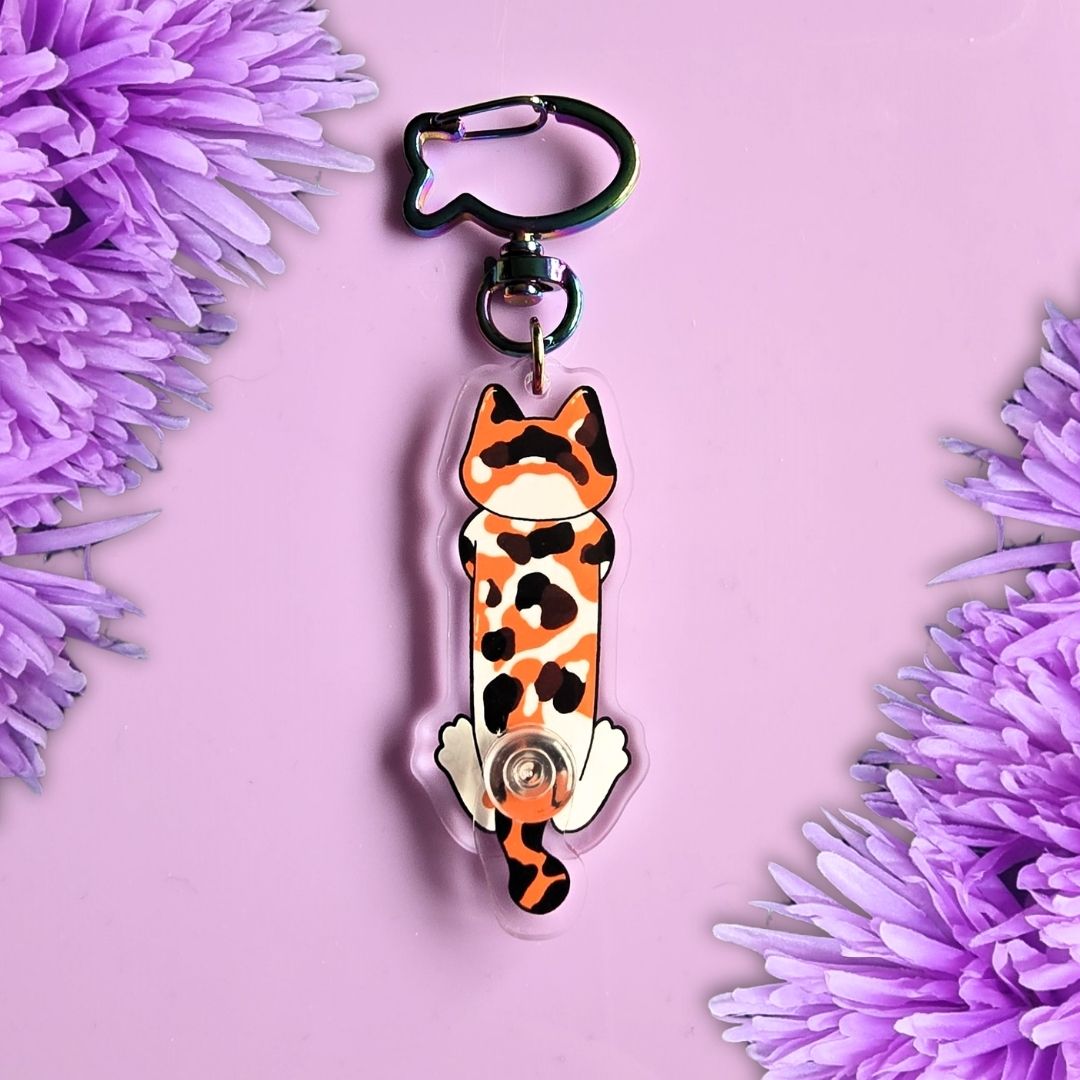 A keychain featuring a stylized cat figure with an orange, black, and white spotted pattern, attached to a black cat-shaped clip, set against a pink background with purple flowers on the sides.
